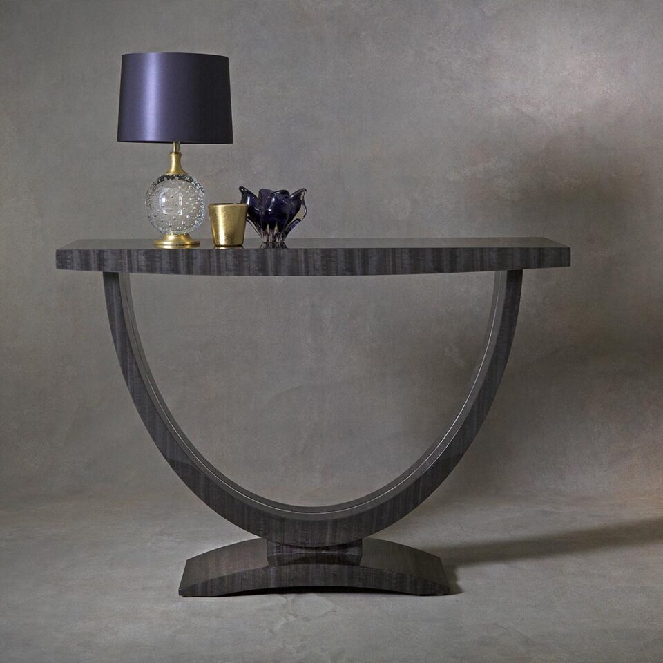 A sleek console table with a wide, curve base finished in a high gloss figured eucalyptus tinted in a smart shade of grey with a polished nickel inlay detail to the base. Design by DAVIDSON London