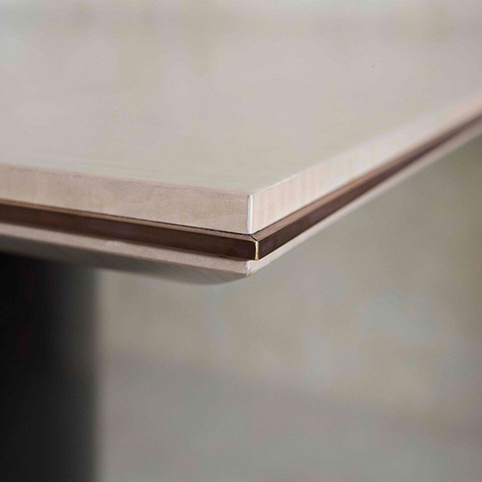 A detail corner image of this contemporary dining table. The tabletop is finished in a satin sycamore silk with an antiqued brushed brass trim. The top stands on three asymmetrical high gloss sycamore black pedestals.